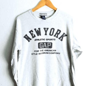 Könnte beinhalten: Ein hellgraues Langarmshirt mit Rundhalsausschnitt und dunkelgrauem Aufdruck auf der Vorderseite: NEW YORK ATHLETIC SPORTS GAP FOR THE AMERICAN STYLE MAXIMUM COMPORT. Das Kleidungsstück hängt an einem Holzkleiderbügel vor einer weißen Wand.