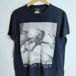 May include: Dark navy blue t-shirt with a black and white graphic print. The graphic features a group of people and the text "WE CUM IN PEACE". The shirt has a crew neck and short sleeves.