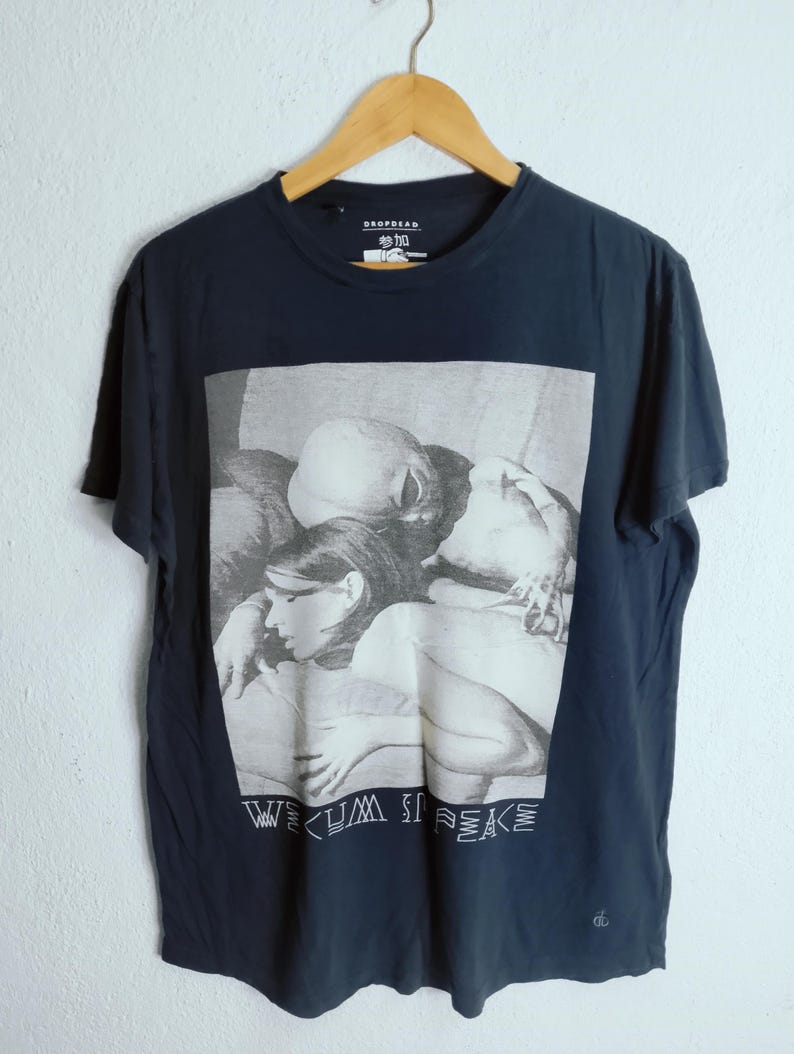 May include: Dark navy blue t-shirt with a black and white graphic print. The graphic features a group of people and the text "WE CUM IN PEACE". The shirt has a crew neck and short sleeves.