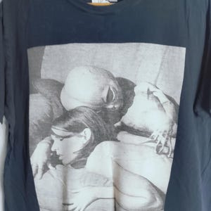 May include: Navy blue t-shirt featuring a black and white image of people. The image has the text "WE CUM IN PEACE" at the bottom. The shirt has a crew neck and short sleeves. The brand name "DROPDEAD" is at the top.