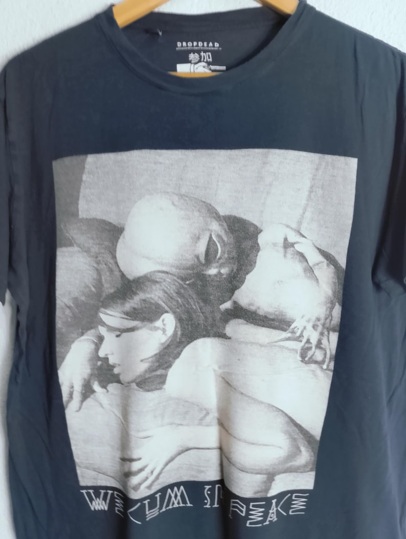 May include: Navy blue t-shirt featuring a black and white image of people. The image has the text "WE CUM IN PEACE" at the bottom. The shirt has a crew neck and short sleeves. The brand name "DROPDEAD" is at the top.
