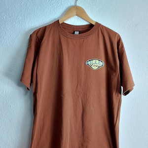 Op de afbeelding: Een bruin T-shirt met een afbeelding van een blauw en geel logo met de tekst "Killer Loop".