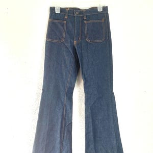 Puede incluir: Pantalones vaqueros acampanados de color azul oscuro con dos bolsillos delanteros grandes y costuras naranjas. Los pantalones cuelgan de una percha blanca sobre un fondo blanco. Las perneras acampanadas son una caracter&iacute;stica destacada.