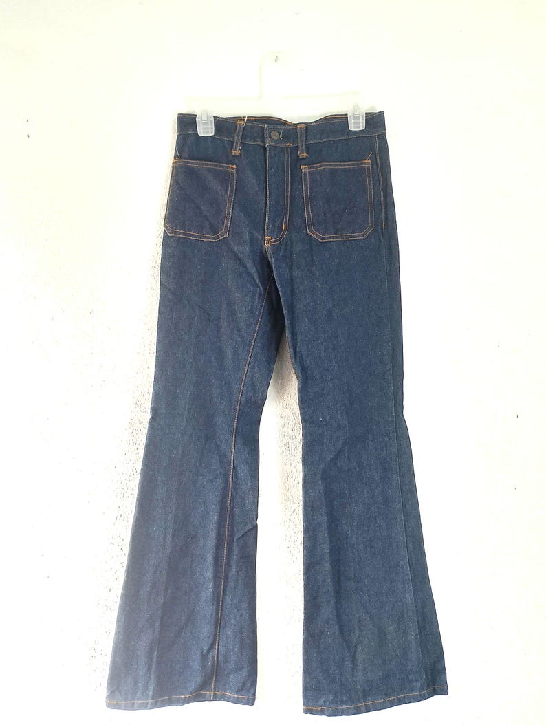 Puede incluir: Pantalones vaqueros acampanados de color azul oscuro con dos bolsillos delanteros grandes y costuras naranjas. Los pantalones cuelgan de una percha blanca sobre un fondo blanco. Las perneras acampanadas son una caracter&iacute;stica destacada.