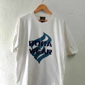 Może przedstawiać: Biała koszulka z nadrukiem niebieskiego i czarnego płomienia oraz napisem "ROCA WEAR" z przodu.