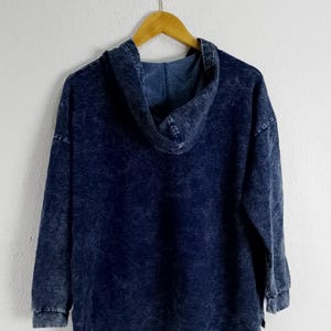 Pode incluir: Um moletom azul marinho com uma apar&ecirc;ncia desbotada e texturizada. O moletom tem capuz, mangas compridas e um ajuste folgado. Est&aacute; pendurado em um cabide de madeira contra uma parede branca.