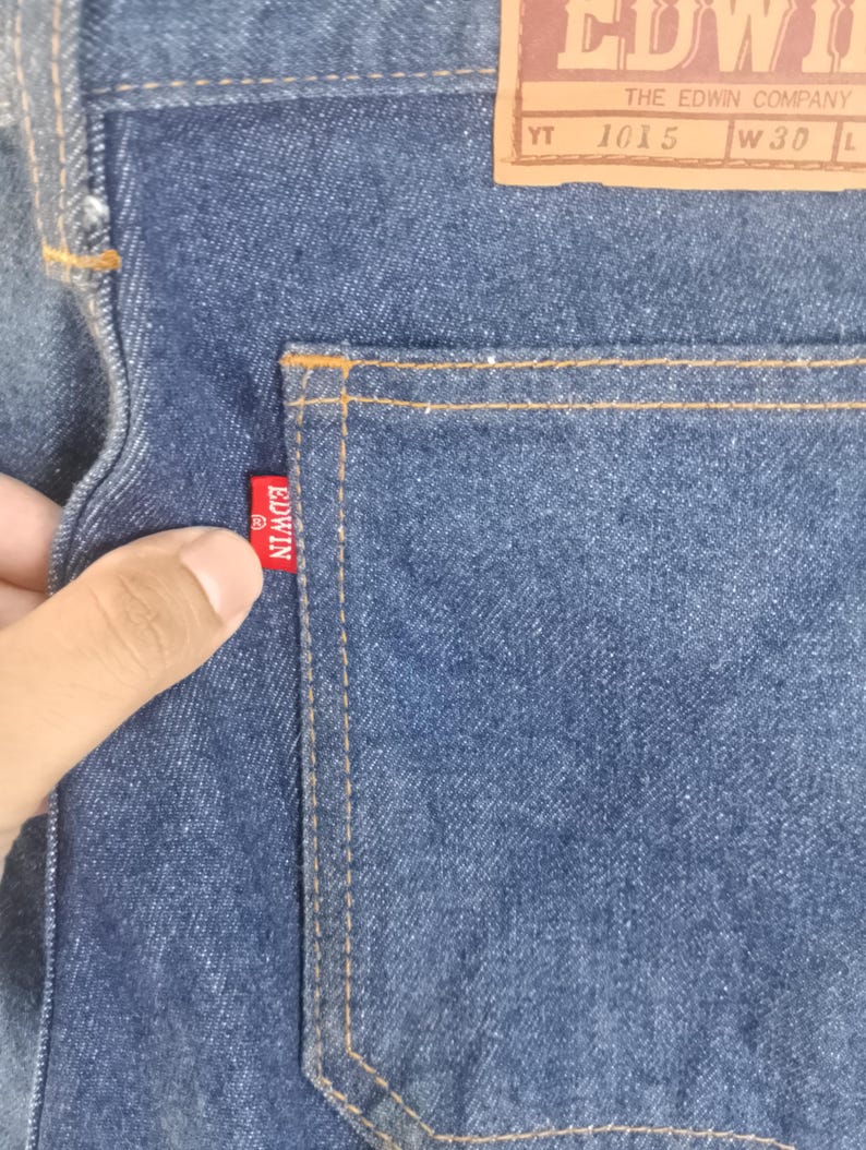 Puede incluir: Primer plano de unos vaqueros de mezclilla azules con un bolsillo trasero y una etiqueta roja de Edwin. La etiqueta Edwin tambi&eacute;n es visible en la parte trasera de los vaqueros. Los vaqueros tienen una etiqueta de cuero marr&oacute;n con el texto "EDWIN THE EDWIN COMPANY 1015 W30".