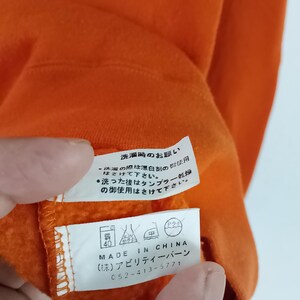 以下が含まれることがあります： オレンジ色の衣類タグのクローズアップ。日本語のテキストとケアシンボルが記載されています。タグには「MADE IN CHINA」と記載されており、会社名と電話番号が含まれています。衣類は鮮やかなオレンジ色です。