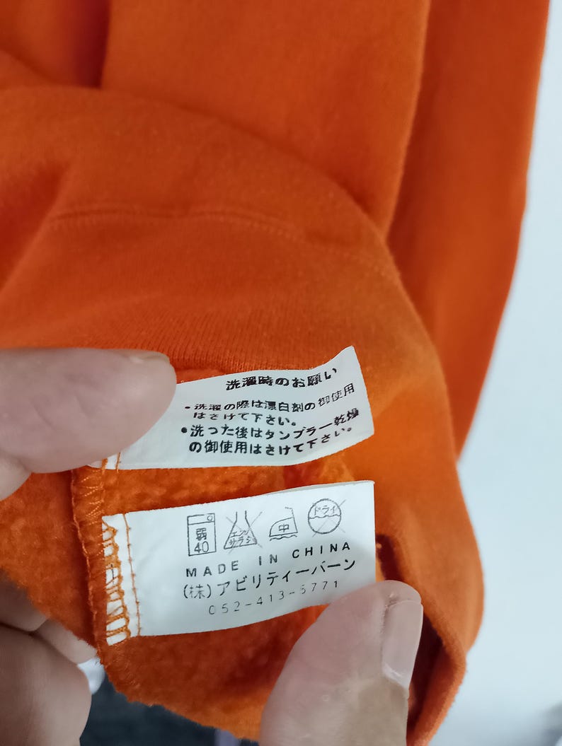 以下が含まれることがあります： オレンジ色の衣類タグのクローズアップ。日本語のテキストとケアシンボルが記載されています。タグには「MADE IN CHINA」と記載されており、会社名と電話番号が含まれています。衣類は鮮やかなオレンジ色です。