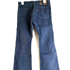 Può includere: Jeans blu scuro con gamba a zampa. I jeans hanno due tasche posteriori, un'etichetta rossa e una toppa in pelle marrone. I jeans sono appesi a una gruccia bianca.