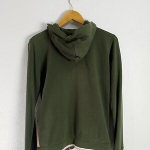 May include: Olive green hoodie with a drawstring hood and a full-length zipper. The hoodie has a white and light pink trim at the bottom and on the cuffs. The back of the hoodie is visible, hanging on a wooden hanger.