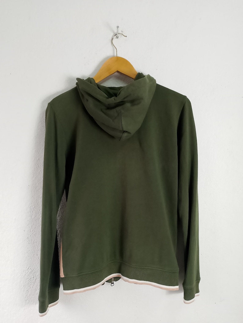 May include: Olive green hoodie with a drawstring hood and a full-length zipper. The hoodie has a white and light pink trim at the bottom and on the cuffs. The back of the hoodie is visible, hanging on a wooden hanger.