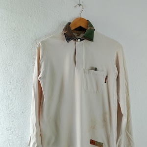 May include: A long-sleeved white polo shirt with a camouflage fabric collar and a small pocket on the left chest. The shirt has a button closure and the text "STUSSY" is visible on the bottom right side of the shirt.