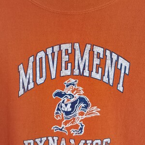 以下が含まれることがあります： オレンジ色のスウェットシャツ。白のブロック体で「MOVEMENT」と「DYNAMICS」の文字がプリントされています。「MOVEMENT」の下には、ネイビーブルーと白の制服を着た漫画の鳥が描かれています。スウェットシャツはクルーネックです。