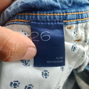 Pode incluir: Close-up de uma etiqueta de roupa de ganga azul. A etiqueta azul marinho exibe o n&uacute;mero "26" em branco. Abaixo, as palavras "SLIM BOYFRIEND" s&atilde;o impressas em branco. O forro interior da roupa &eacute; vis&iacute;vel.