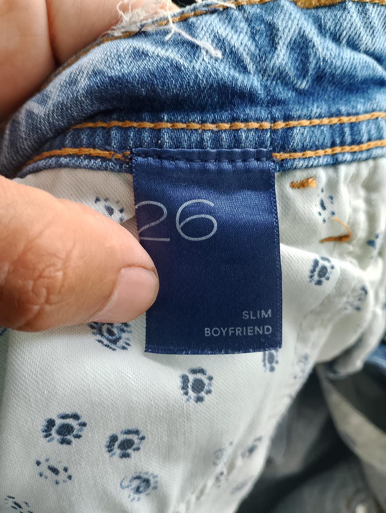 Pode incluir: Close-up de uma etiqueta de roupa de ganga azul. A etiqueta azul marinho exibe o n&uacute;mero "26" em branco. Abaixo, as palavras "SLIM BOYFRIEND" s&atilde;o impressas em branco. O forro interior da roupa &eacute; vis&iacute;vel.