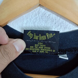 Puede incluir: Primer plano de una etiqueta de prenda negra con el texto "Keep Your Ocean Blue." La etiqueta tambi&eacute;n incluye el texto "USA BODY GLOVE AND THE HAND DESIGN ARE REGISTERED TRADEMARKS OF DIVE N' SURF, INC."