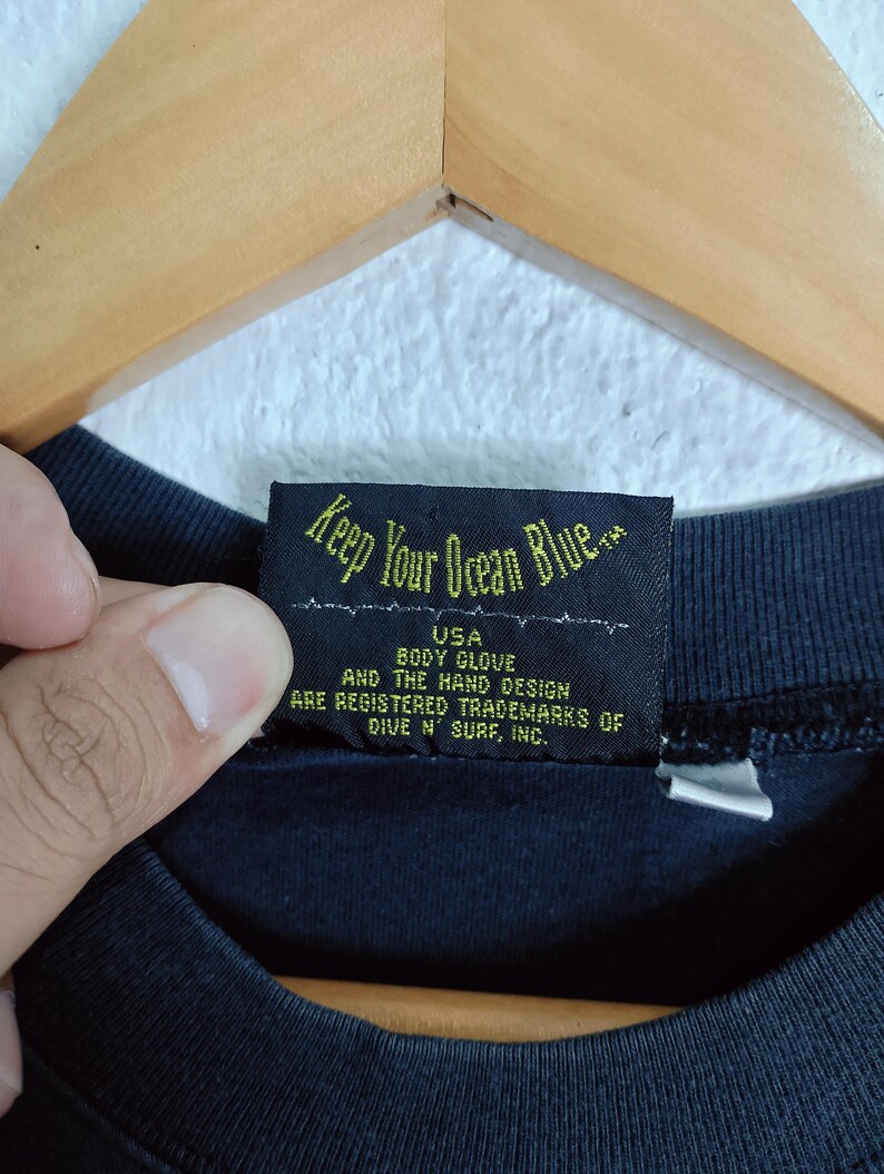 Puede incluir: Primer plano de una etiqueta de prenda negra con el texto "Keep Your Ocean Blue." La etiqueta tambi&eacute;n incluye el texto "USA BODY GLOVE AND THE HAND DESIGN ARE REGISTERED TRADEMARKS OF DIVE N' SURF, INC."