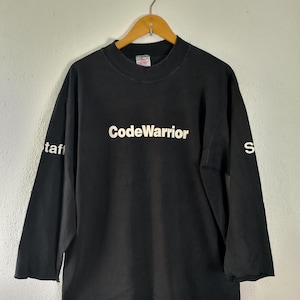 Puede incluir: Una camiseta negra de manga larga con cuello alto. La palabra CodeWarrior está impresa en blanco en el centro del pecho. La palabra staff está impresa en blanco en la manga derecha y la letra S está impresa en blanco en la manga izquierda.