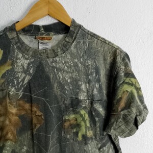 May include: Camouflage pattern t-shirt with a chest pocket. The shirt features a green, brown, and gray woodland camouflage design. The shirt is hanging on a wooden hanger.