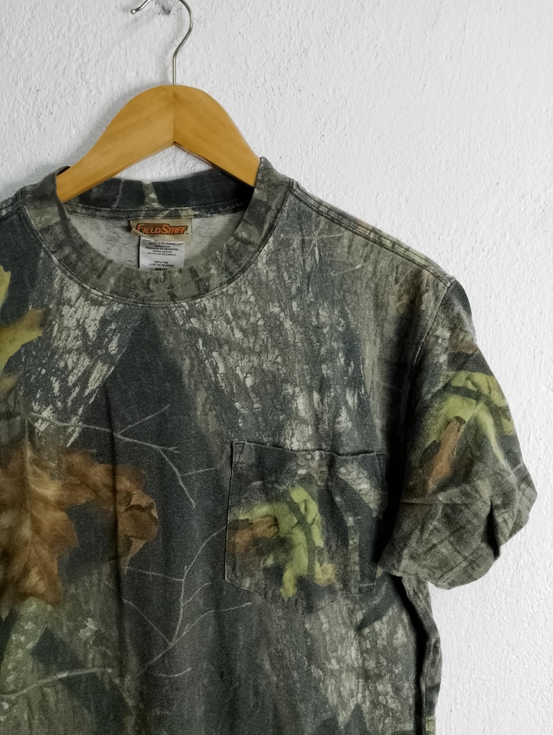 May include: Camouflage pattern t-shirt with a chest pocket. The shirt features a green, brown, and gray woodland camouflage design. The shirt is hanging on a wooden hanger.