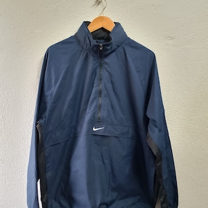 Puede incluir: Una chaqueta cortavientos azul marino con un logotipo Nike Swoosh blanco en el pecho. La chaqueta tiene una cremallera de media cremallera y un cordón en la parte inferior.