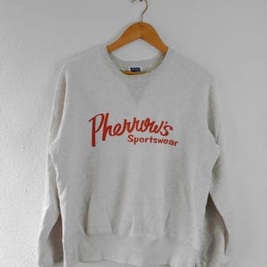 Może przedstawiać: Jasnoszara bluza z czerwonym napisem "Pherrow's Sportswear" na piersi. Bluza wisi na drewnianym wieszaku na tle białej ściany. Ma okrągły dekolt i długie rękawy.