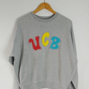 Puede incluir: Sudadera gris con las letras "UCB" en rojo, amarillo y azul cosidas en la parte delantera. La sudadera está colgada en una percha de madera, sobre un fondo claro.