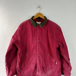 Puede incluir: Chaqueta burdeos con cuello verde y dos bolsillos delanteros. La chaqueta tiene cierre de botones y está colgada de una percha de madera. La chaqueta está hecha de un material duradero.