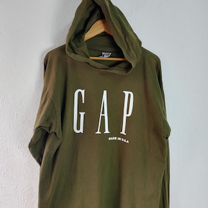 May include: An olive green hooded sweatshirt with the white text "GAP" across the chest. Below the text is the phrase "MADE IN U.S.A." The sweatshirt is hanging on a wooden hanger.