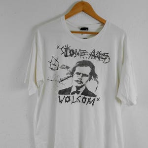 May include: White t-shirt with a black and white graphic of a man's portrait and the text "STONE AGE" and "VOLCOM". The shirt is hanging on a wooden hanger.