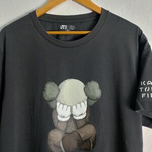 Puede incluir: Camiseta gris oscuro con un gr&aacute;fico de un personaje de dibujos animados que se cubre los ojos. El personaje est&aacute; en tonos de marr&oacute;n, gris y blanco. La camiseta tiene el texto "KA TO FI" en la manga y "UT" con texto japon&eacute;s en el pecho.