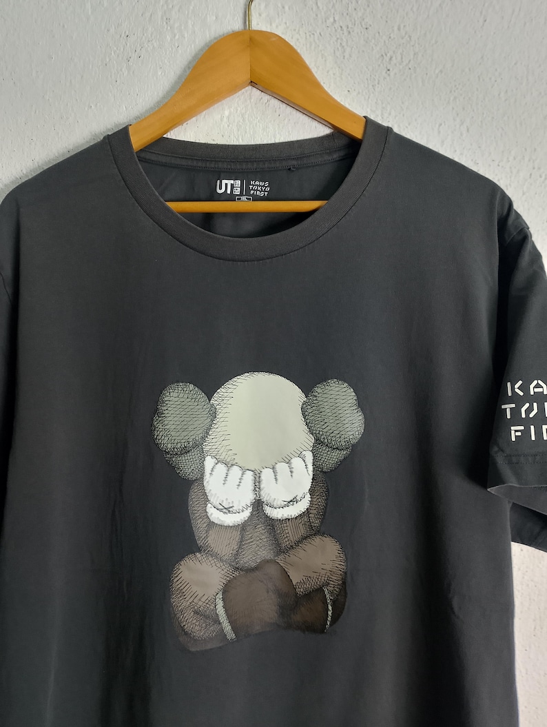Puede incluir: Camiseta gris oscuro con un gr&aacute;fico de un personaje de dibujos animados que se cubre los ojos. El personaje est&aacute; en tonos de marr&oacute;n, gris y blanco. La camiseta tiene el texto "KA TO FI" en la manga y "UT" con texto japon&eacute;s en el pecho.