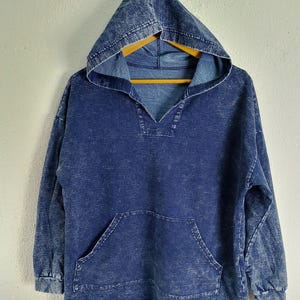 Pode incluir: Um moletom azul jeans com bolso frontal e capuz. O moletom tem decote em V e mangas compridas. O jeans tem uma apar&ecirc;ncia desbotada e texturizada.
