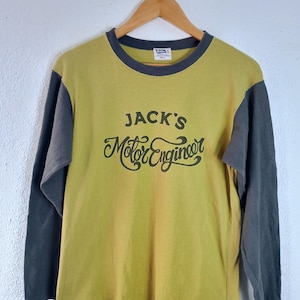 Peut inclure: T-shirt à manches longues jaune moutarde avec des manches et un col gris. Le t-shirt porte l'inscription "JACK'S Motor Engineer" dans une police vintage. Le t-shirt est suspendu à un cintre en bois sur fond blanc.