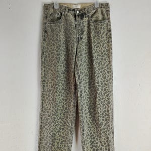 May include: A pair of straight-leg jeans with a leopard print pattern in shades of gray and beige. The jeans have a button fly and classic five-pocket design. They are hanging on a white hanger against a white wall.