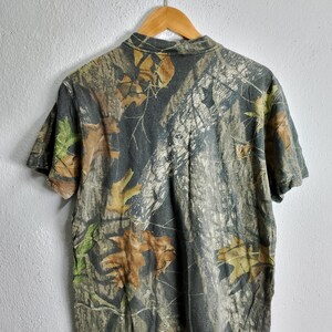 May include: Short-sleeved camouflage t-shirt with a collared neckline. The shirt features a pattern of brown, green, and tan leaves and branches on a dark gray background. The shirt is hanging on a wooden hanger.