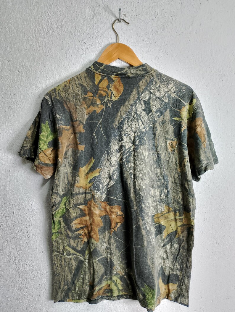 May include: Short-sleeved camouflage t-shirt with a collared neckline. The shirt features a pattern of brown, green, and tan leaves and branches on a dark gray background. The shirt is hanging on a wooden hanger.