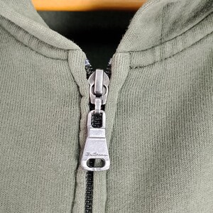 May include: Close-up of an olive green hooded sweatshirt with a silver zipper. The zipper pull has the brand name "Ben Sherman" engraved on it. The fabric appears to be a soft, knit material.