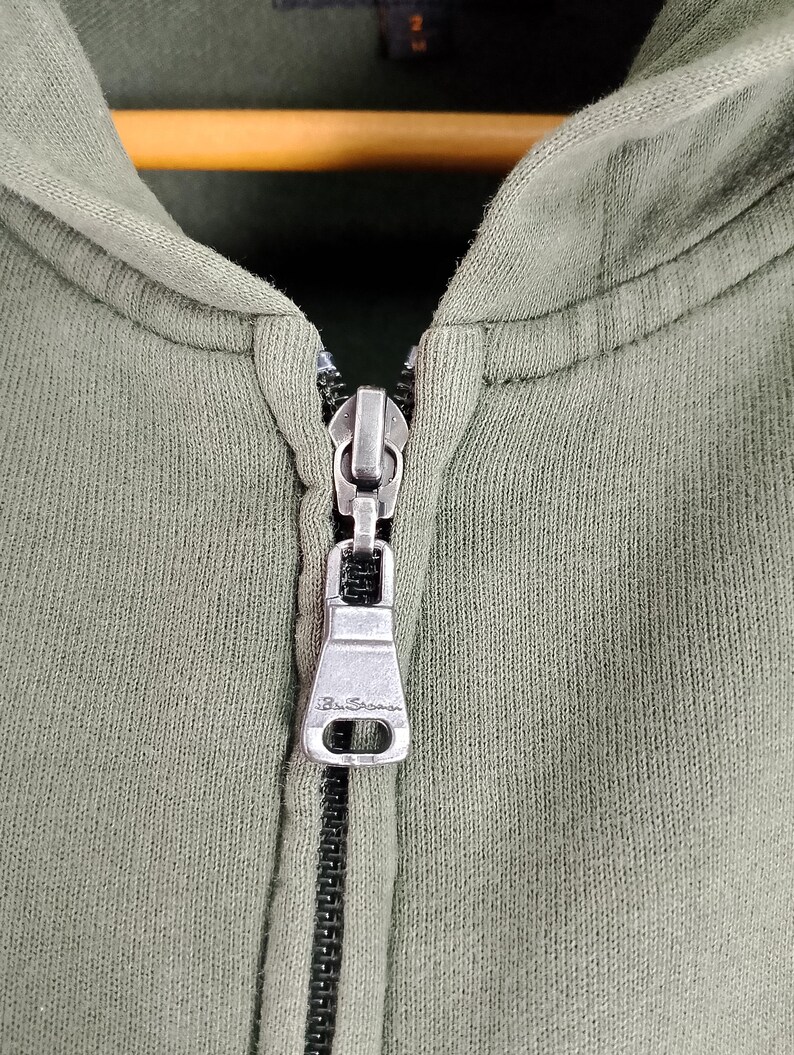 May include: Close-up of an olive green hooded sweatshirt with a silver zipper. The zipper pull has the brand name "Ben Sherman" engraved on it. The fabric appears to be a soft, knit material.