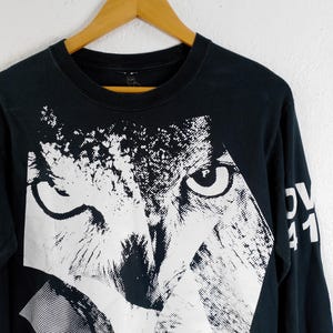 Pode incluir: Camiseta preta de manga comprida com um grande gr&aacute;fico branco do rosto de um animal. O design &eacute; uma representa&ccedil;&atilde;o estilizada e geom&eacute;trica com olhos vis&iacute;veis e uma apar&ecirc;ncia texturizada. A manga tem o texto "OV 41" em branco.