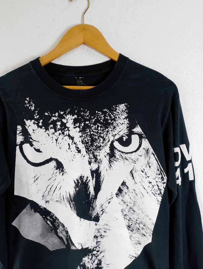 Pode incluir: Camiseta preta de manga comprida com um grande gr&aacute;fico branco do rosto de um animal. O design &eacute; uma representa&ccedil;&atilde;o estilizada e geom&eacute;trica com olhos vis&iacute;veis e uma apar&ecirc;ncia texturizada. A manga tem o texto "OV 41" em branco.