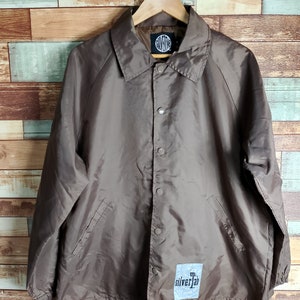 May include: A brown nylon coach jacket with a snap closure and a Silvertab logo patch on the left chest. The jacket has a drawstring at the bottom and two pockets on the front.