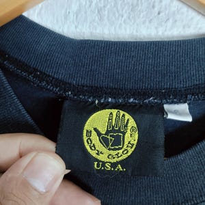 Puede incluir: Primer plano de una camiseta azul oscuro con una etiqueta negra. La etiqueta presenta un c&iacute;rculo amarillo con un logotipo de mano y el texto "BODY GLOVE U.S.A."