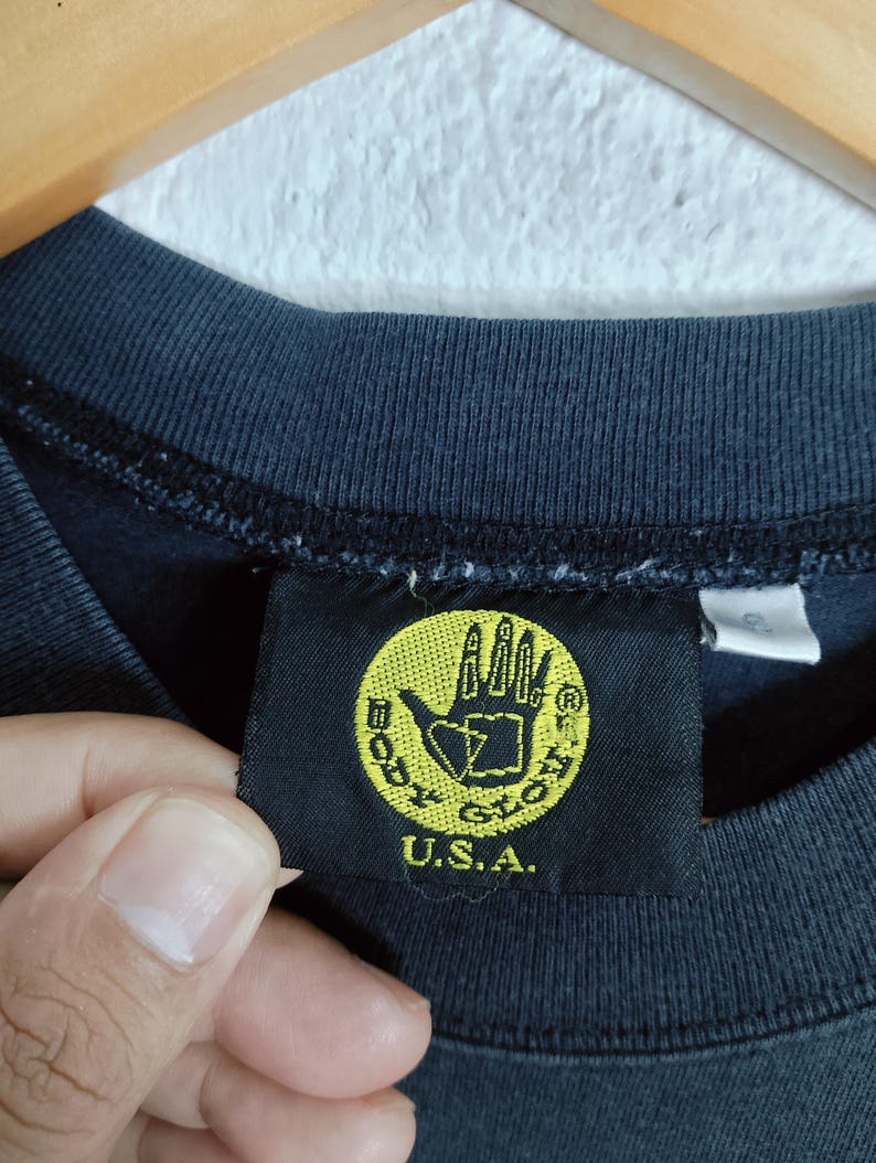 Puede incluir: Primer plano de una camiseta azul oscuro con una etiqueta negra. La etiqueta presenta un c&iacute;rculo amarillo con un logotipo de mano y el texto "BODY GLOVE U.S.A."