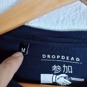 May include: Close-up of a navy blue t-shirt with the brand name "DROP DEAD" and Japanese characters. The shirt has a size "M" tag. A hand is pointing at the size tag.