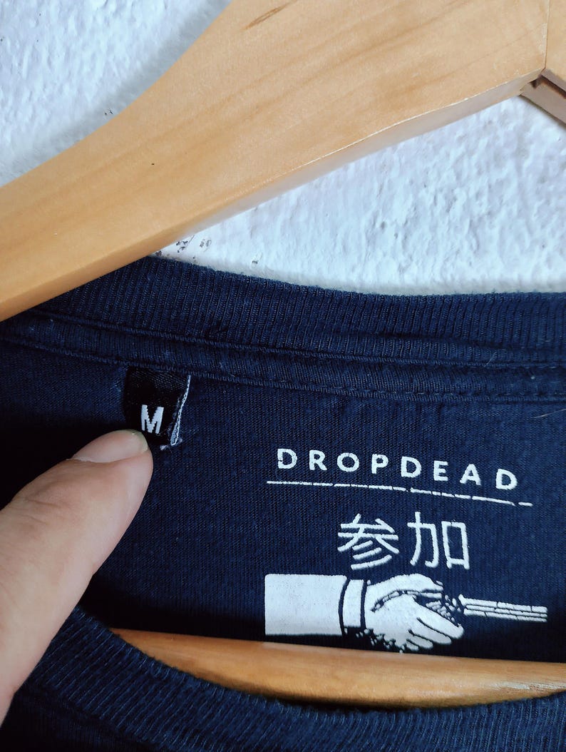 May include: Close-up of a navy blue t-shirt with the brand name "DROP DEAD" and Japanese characters. The shirt has a size "M" tag. A hand is pointing at the size tag.