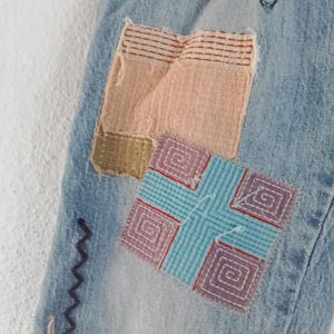 Pode incluir: Close-up de jeans de ganga azul claro com patches coloridos. Os patches incluem ret&acirc;ngulos tecidos em rosa, bege e listras vermelhas, e um quadrado com uma cruz azul e um design de espiral roxa. Costura decorativa em roxo e branco.