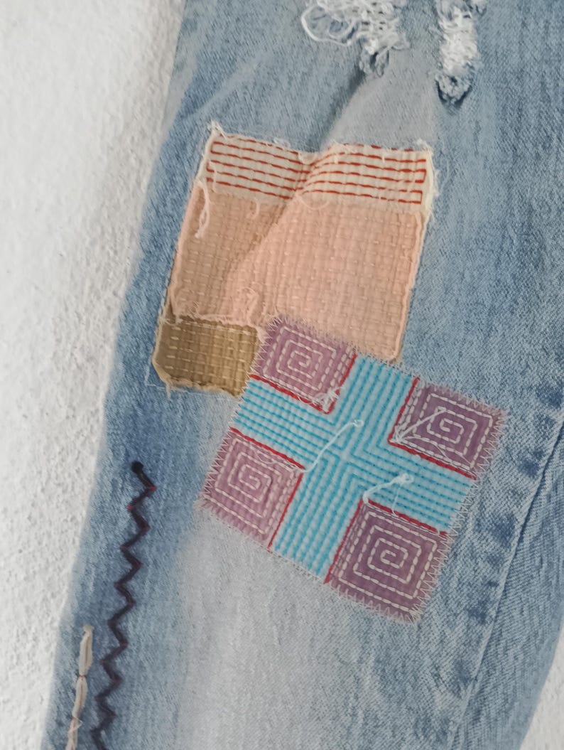 Pode incluir: Close-up de jeans de ganga azul claro com patches coloridos. Os patches incluem ret&acirc;ngulos tecidos em rosa, bege e listras vermelhas, e um quadrado com uma cruz azul e um design de espiral roxa. Costura decorativa em roxo e branco.