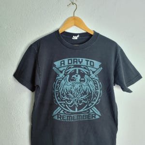 Puede incluir: Una camiseta negra de manga corta con un estampado gráfico azul claro de un águila centrada en un emblema circular, flanqueada por dos espadas cruzadas. El texto A DAY TO REMEMBER está impreso en una fuente de bloque sobre y bajo el emblema.